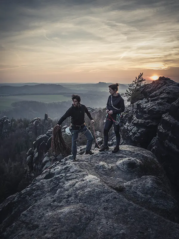 Twee klimmers met hun uitrusting staan bij zonsondergang op een rotsachtige klif en kijken uit over een uitgestrekt landschap van heuvels, bossen en bergen in de verte, onder een bewolkte hemel.